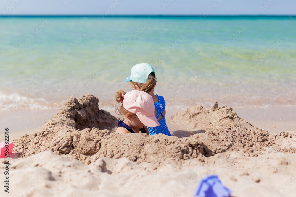 Two sister girls in swimsuits and caps playing on the beach with toys and sand on a sunny summer day. Fun family vacation at the sea. Activity outdoors