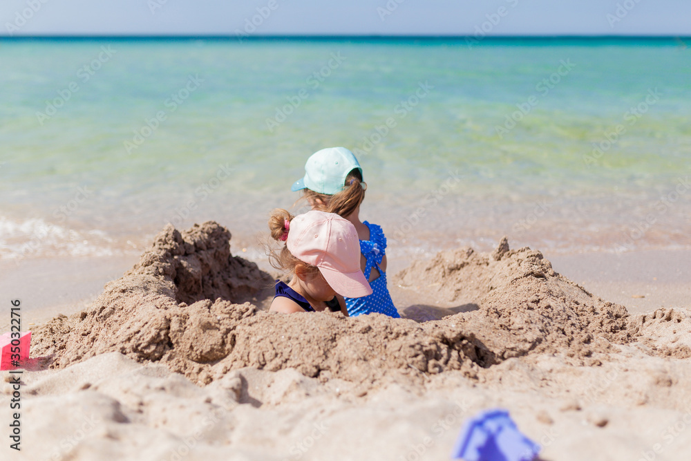 Two sister girls in swimsuits and caps playing on the beach with toys and sand on a sunny summer day. Fun family vacation at the sea. Activity outdoors Two sister girls in swimsuits and caps playing on the beach with toys and sand on a sunny summer day. Fun family vacation at the sea. Activity outdoors