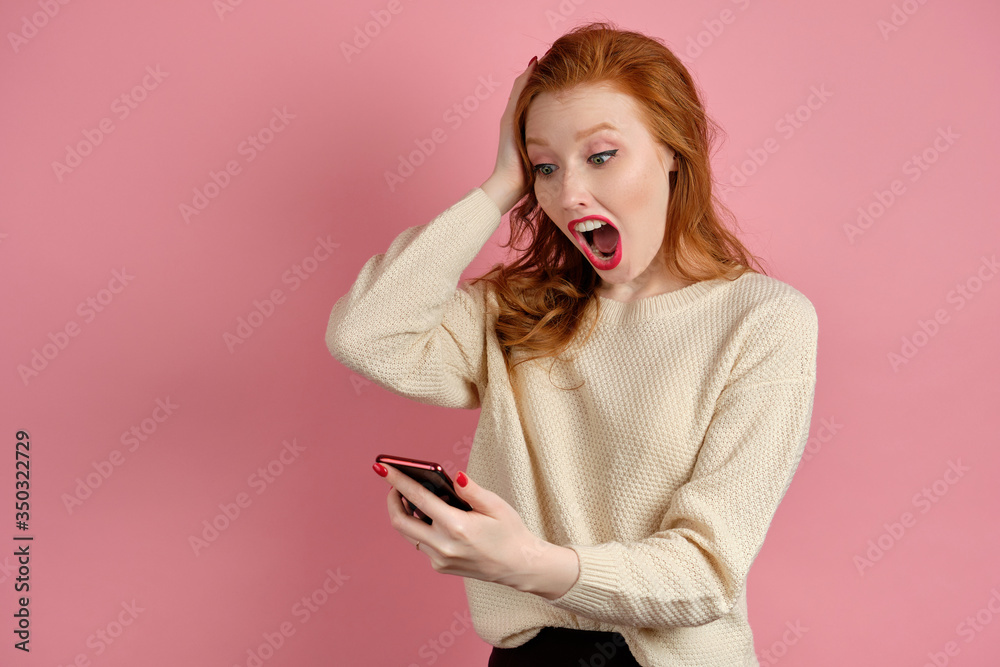 A red-haired girl with red lipstick stands in a pink background and looks at the phone in hand shocked by opening mouth and holding hand on head A red-haired girl with red lipstick stands in a pink background and looks at the phone in hand shocked by opening mouth and holding hand on head