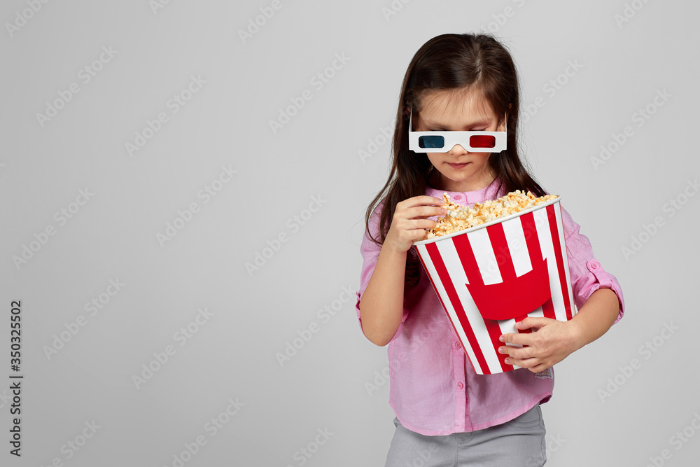 cute child girl wearing red-blue 3d glasses and eating popcorn from bucket isolated on gray background. copy space cute child girl wearing red-blue 3d glasses and eating popcorn from bucket isolated on gray background. copy space