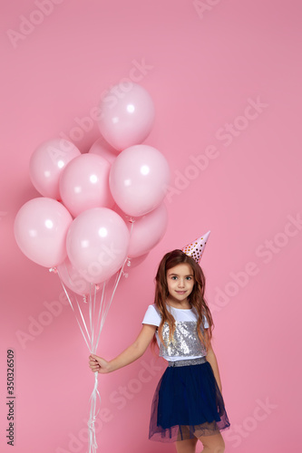 Beautiful happy little child girl in dress and birthday hat celebrating with pastel pink air balloons isolated on pink background. birthday party.