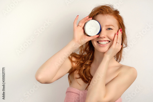 Beautiful red-haired girl with clean skin stands on a white background, smiling hiding behind a can of cream.