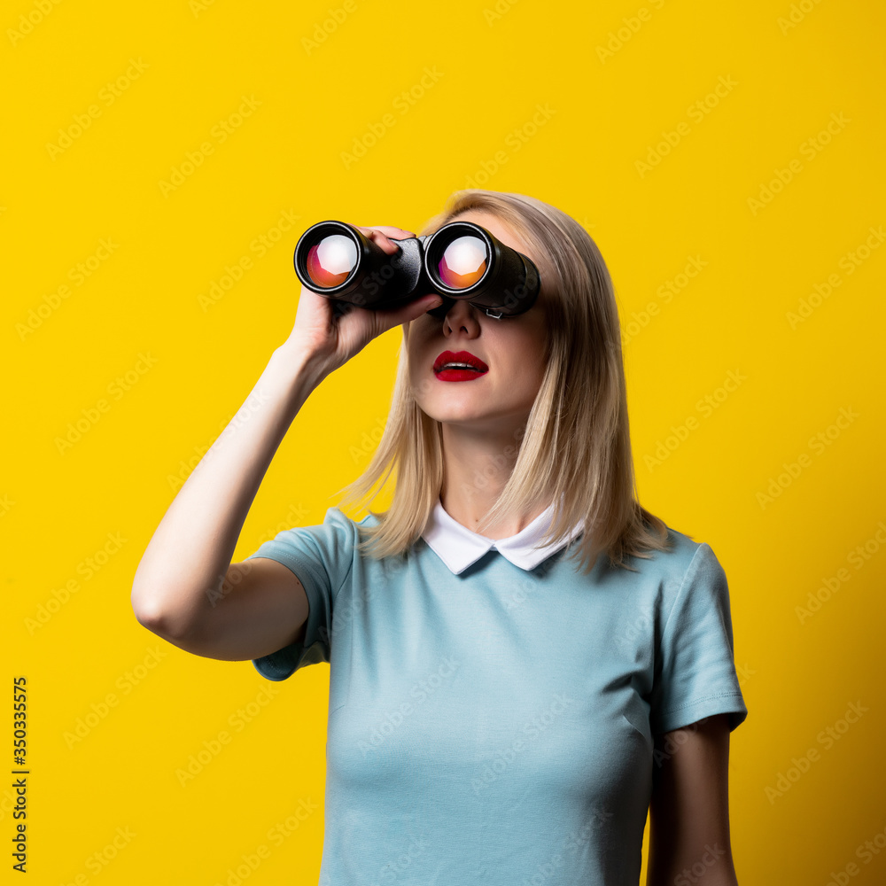 Blonde girl in blue dress watching in binoculars on yellow background Blonde girl in blue dress watching in binoculars on yellow background