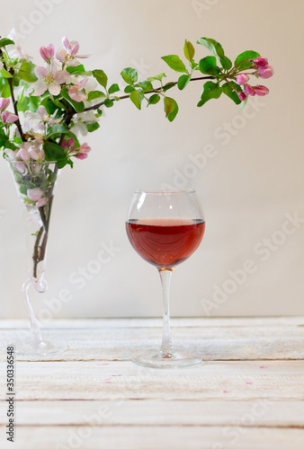Glass of pink wine and apple tree branches with pink flowers in a vase on white wooden table