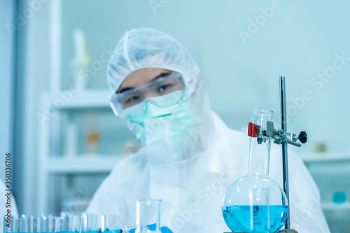 two scientists wearing protective glasses  white jumpsuit  and medical face mask  analysing  biological sample in test tube glassware in science laboratory with microscope for virus pandemic