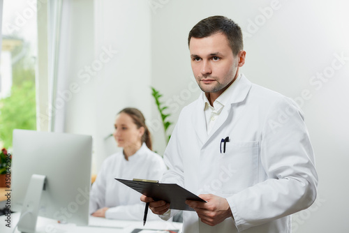 A serious surgeon with bristles in a white coat holds a black clipboard with documents and a pen in a hospital. A female doctor sitting in front of the desktop computer in the corner of the room. 