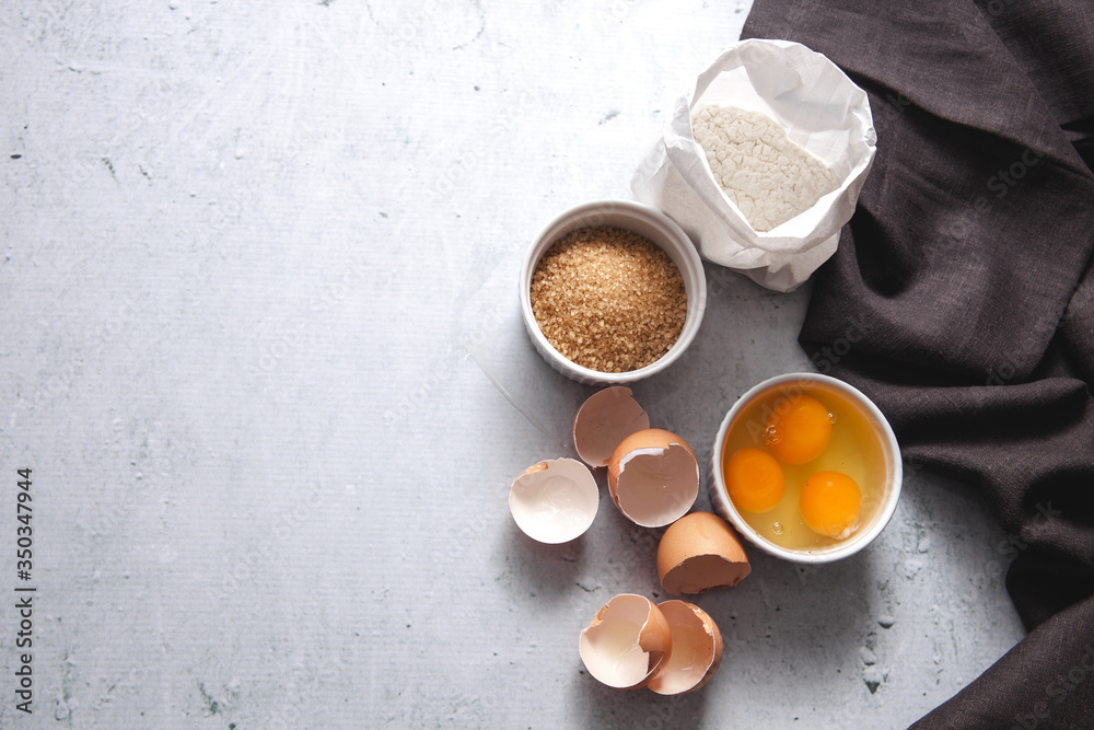 Dough baking ingredients: a bag of flour, eggs, egg with yolk in a bowl, brown sugar with rolling pin on light grey concrete background. Top view, flatlay, copy space. Recipe Mockup