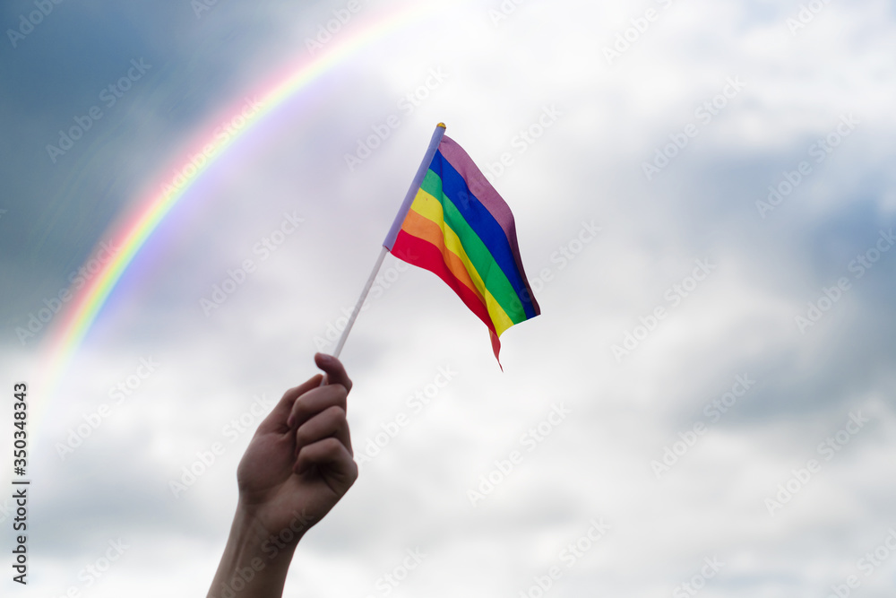 A hand holds an LGBT flag against a sky with clouds and a rainbow. A hand holds an LGBT flag against a sky with clouds and a rainbow.
