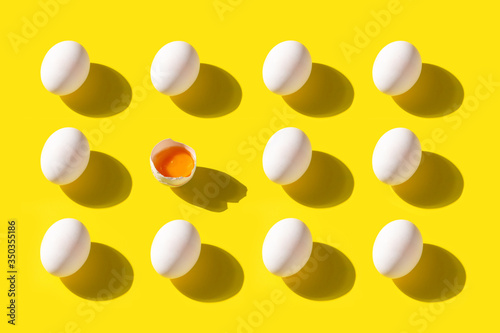 Pattern of white chicken eggs on a yellow background with a shadow. One egg is broken. Poultry products.