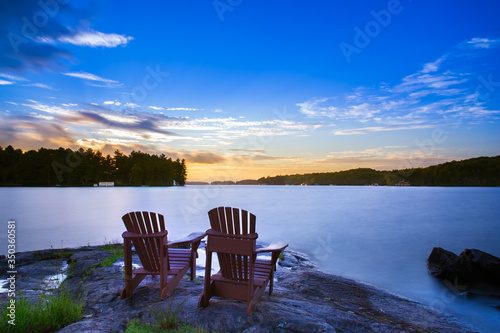 Long exposure of two Muskoka chairs sitting on a rock formation facing a calm lake at dusk in cottage country.
