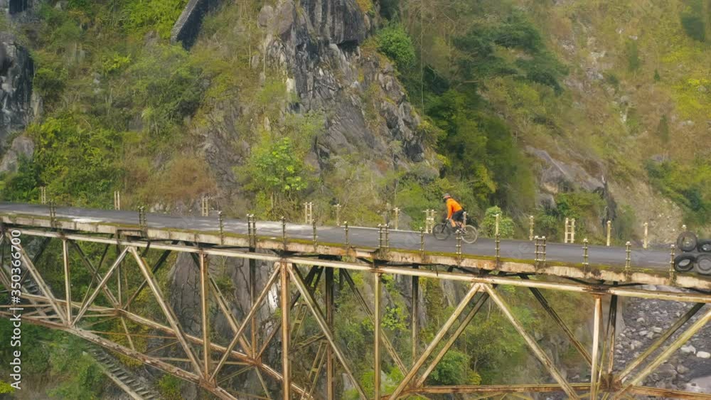 Drone view of man on bicycle riding on suspension bridge in mountains Drone view of man on bicycle riding on suspension bridge in mountains