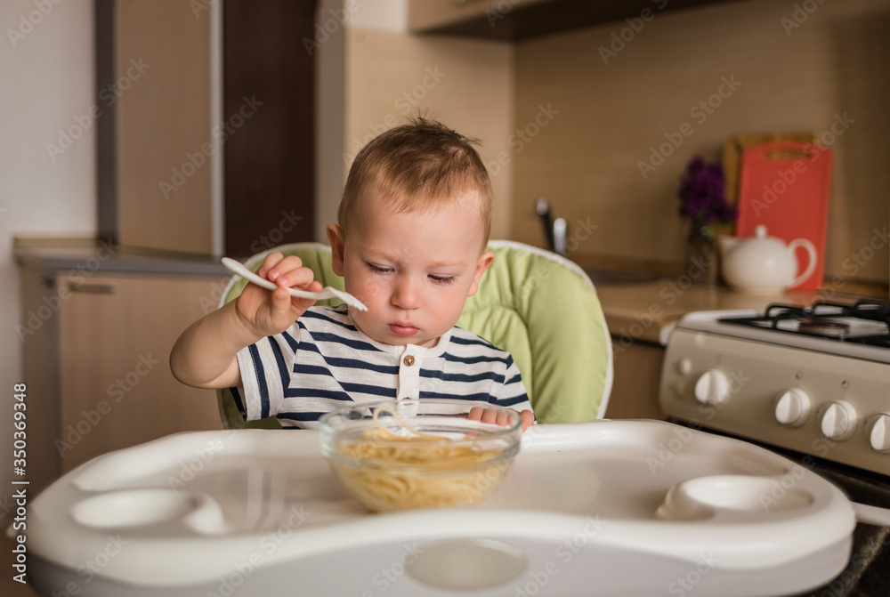 Cute little boy learn to eat by himself with a fork. Baby eats spaghetti sitting on a high chair in the kitchen. Cute little boy learn to eat by himself with a fork. Baby eats spaghetti sitting on a high chair in the kitchen.