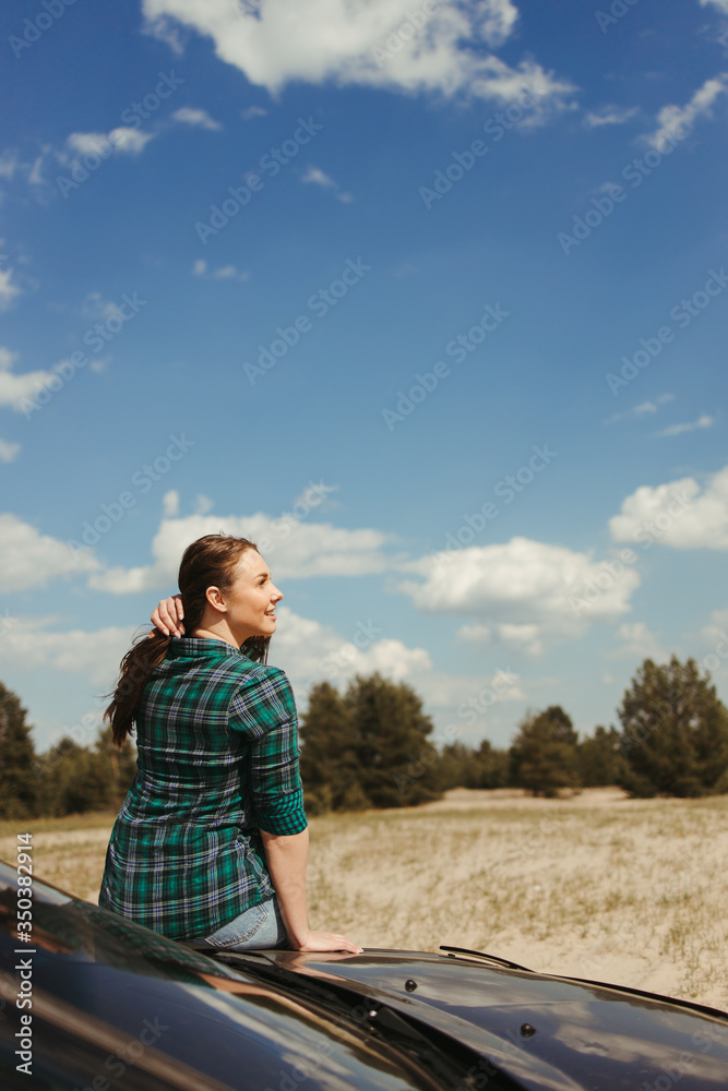 Young woman traveler sitting on the car hood while traveling. Road trip, vacation and adventures
