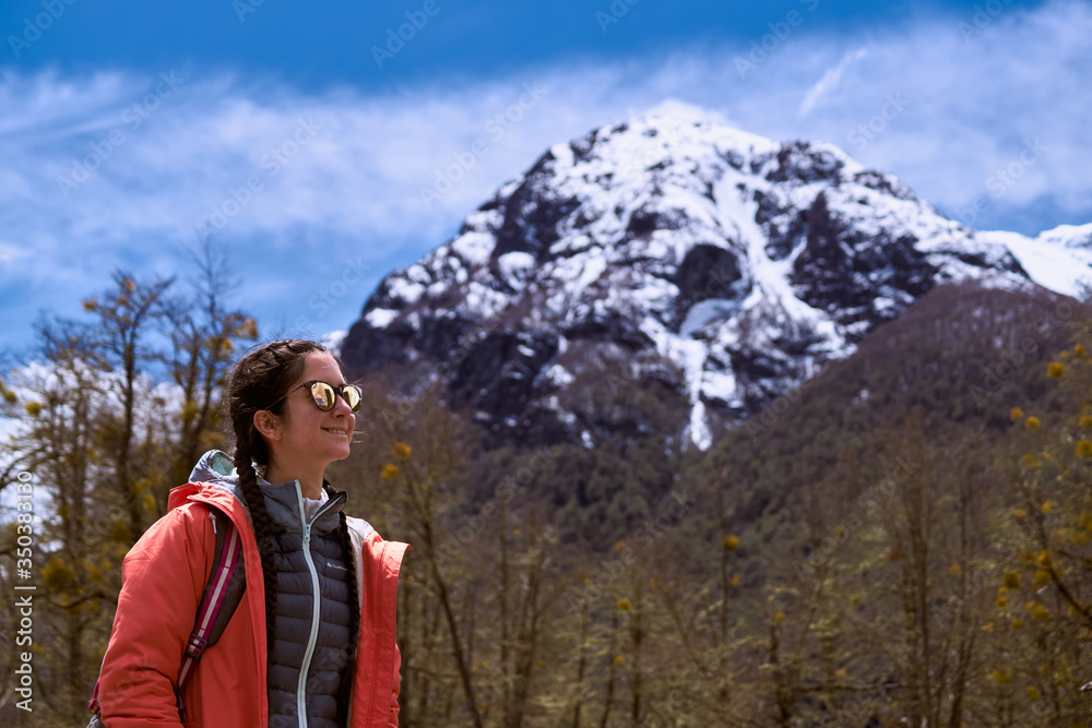 Beautiful and colorful portrait in a cold winter of a smiling beautiful young woman wearing orange jacket and sunglasses. Surrounded by a dry forest with snow capped mountains behind and a blue sky