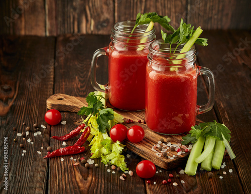 Two glasses with fresh tomato juice, celery, parsley and ripe tomatoes on dark brown wooden background.