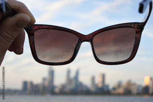 POV of person holding sunglasses on Swan River riverbank looking at the landscape view of Perth city financial centre skyline at sunset