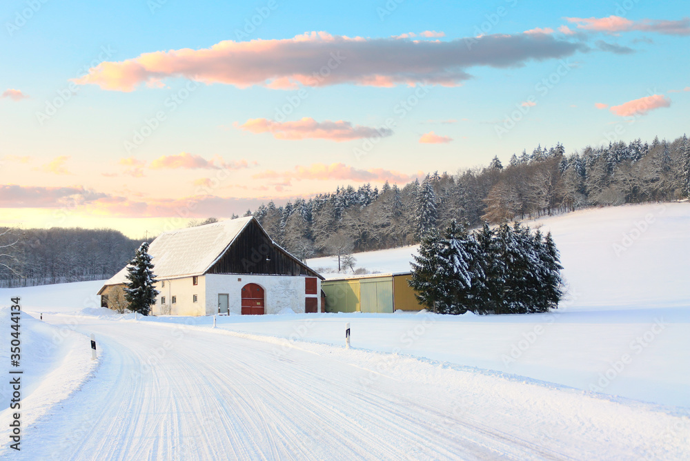 A snowy winter landscape in East Westphalia in Germany. In the middle a farmhouse and in the background a fir forest. In the foreground is a curve with a white, snow-covered road.