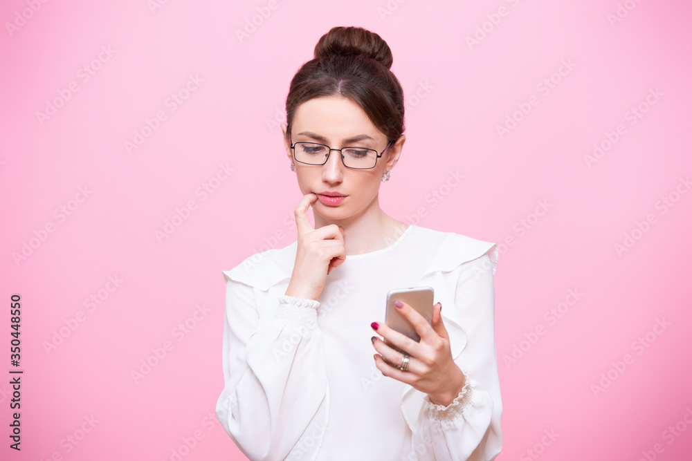 A girl in a business suit with a mobile phone in her hands, thoughtfully looks at the screen. Isolated on a pink background A girl in a business suit with a mobile phone in her hands, thoughtfully looks at the screen. Isolated on a pink background