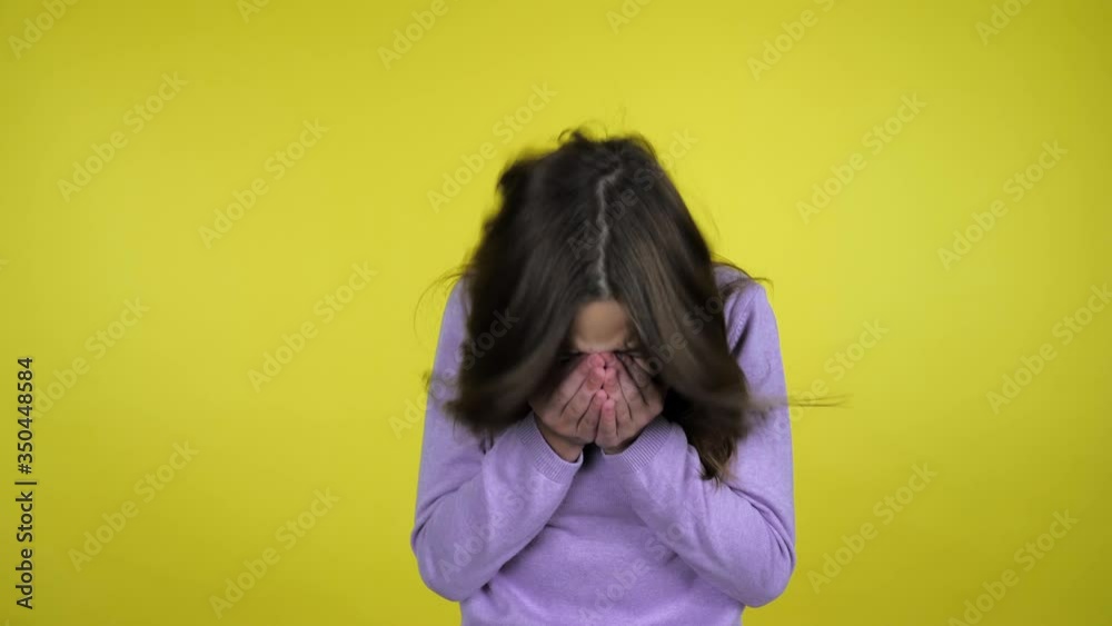 Teen girl in a pink sweater sneezes covering her face with palms on a yellow background with copy space. Place for text or product. 4K slow motion footage