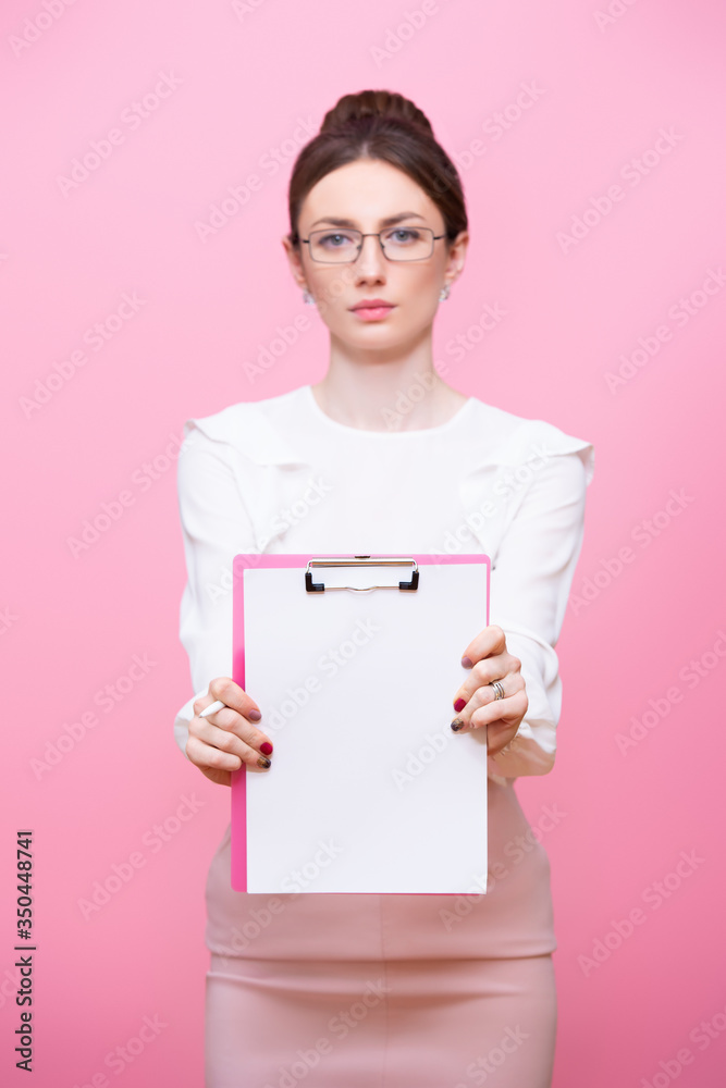 A young woman with glasses holds a clipboard folder in her hands with a serious expression on her face. Isolated on pink background A young woman with glasses holds a clipboard folder in her hands with a serious expression on her face. Isolated on pink background
