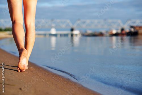 Woman legs and feet walking on the sand of the beach with the sea water in urban city background