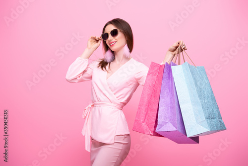Fashionable elegant girl in sunglasses with multi-colored shopping bags is happy from discounts and sale. Isolated on pink background