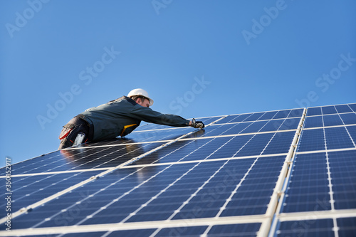 Male worker mounting solar modules, panels and support structures of photovoltaic solar array. Electrician wearing safety helmet and gloves. Concept of sun energy and power sustainable resources.