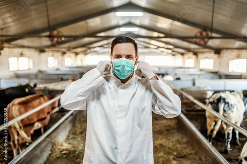 Young Caucasian veterinarian in protective uniform standing in barn and putting on protective mask on face.