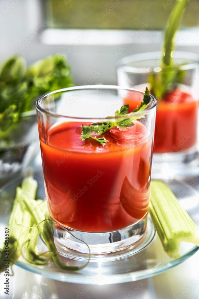 Two glasses of tomato juice with parsley and celery decorations, stand near the window, morning sunlight shines, shallow depth of field, selective focus. Natural drinks concept. Two glasses of tomato juice with parsley and celery decorations, stand near the window, morning sunlight shines, shallow depth of field, selective focus. Natural drinks concept.