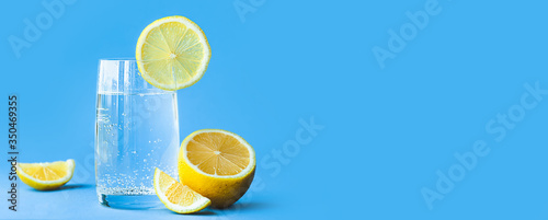translucent glass of water with gases and bubbles, with a round slice of lemon on a glass and a whole lemon on blue background. Refreshment in the heat. Healthy banner lifestyle and nutrition