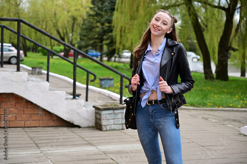 Portrait of a young pretty girl with a smile in a black jacket and jeans. Model posing standing in spring park outdoors in the city.