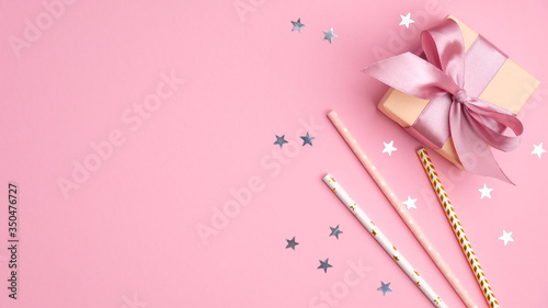 Gift box and drinking straws with confetti stars on pink table top view. Girls birthday, wedding or Christmas party concept.