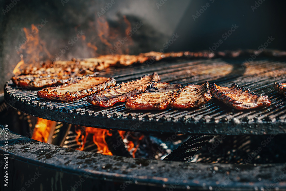 Grilling ribs for barbecue outdoors in summer. Food background with barbecue. Pork ribs cooked on a flaming large round grill with selective focus. Harmful high-calorie foods. Street meat

