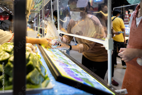 Asian woman in a protective mask,buying food in food court at shopping mall after Coronavirus quarantine or Covid-19 with plastic shield partition,social distancing safety,new normal city lifestyle