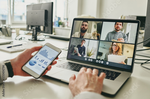 Close-up of businessman working in empty office and using smartphone to view graph while discussing sales with colleagues via video conferencing app photo