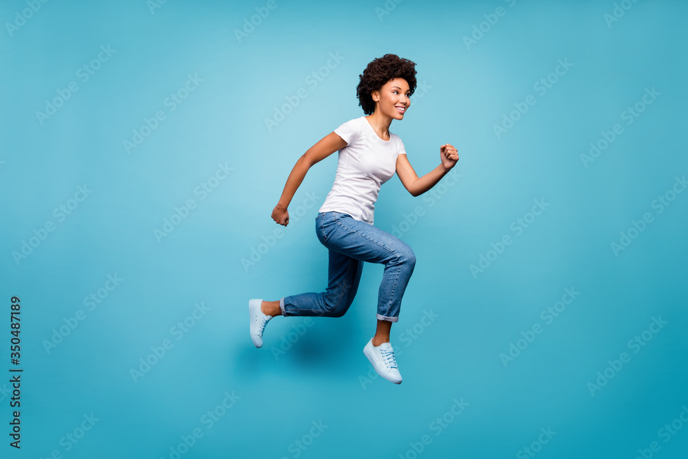 Full length photo of funky dark skin curly lady jumping high sporty person rushing discount shopping final day wear casual white t-shirt jeans isolated blue color background