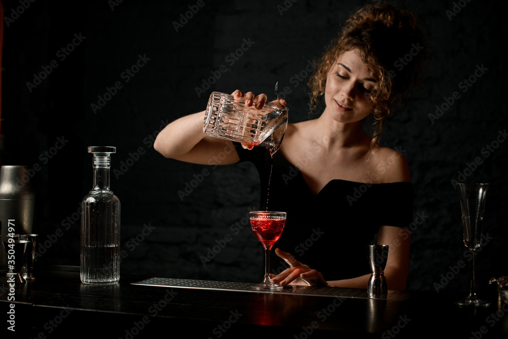 Smiling woman bartender in dark bar pours cocktail from cup to glass