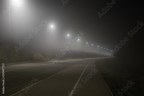 Empty road and sidewalk under white street lights in night. Dark time. Foggy air. Poor visibility.