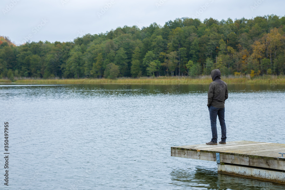 One young man standing alone on edge of wooden footbridge and staring at lake. Hooded guy. Peaceful atmosphere in nature. Enjoying fresh air in cold day. Back view.