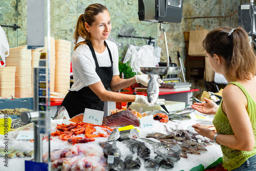 Bild auf Leinwand Portrait of friendly female fishmonger showing raw European bass to woman behind