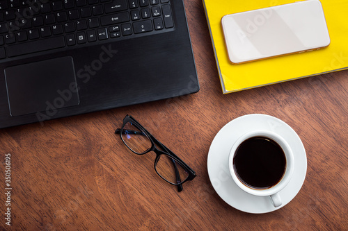 Top view of black coffee with glasses, book, phone and laptop on wooden background.