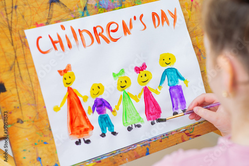Girl is drawing the colorful children by watercolors with words Children's Day on a wooden easel for the holiday Happy Children's Day.