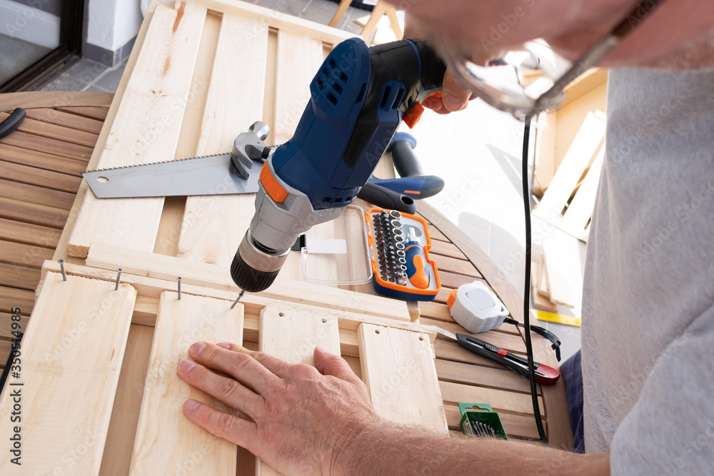 Craftsman using electric screwdriver to fix wooden shelves laying on round table. Handyman working at balcony during self-isolation. House improving and home decoration during quarantine concept Craftsman using electric screwdriver to fix wooden shelves laying on round table. Handyman working at balcony during self-isolation. House improving and home decoration during quarantine concept