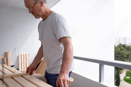 Focused man in eyeglasses and t-shirt repairing shelves at balcony. Senior worker adjusting two parts on round table. House improving, DIY and home decoration during quarantine concept