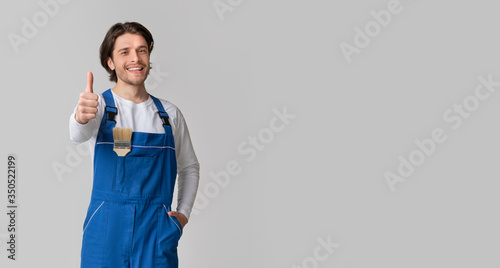 Smiling handyman with paintbrush in pocket showing thumb up, light background