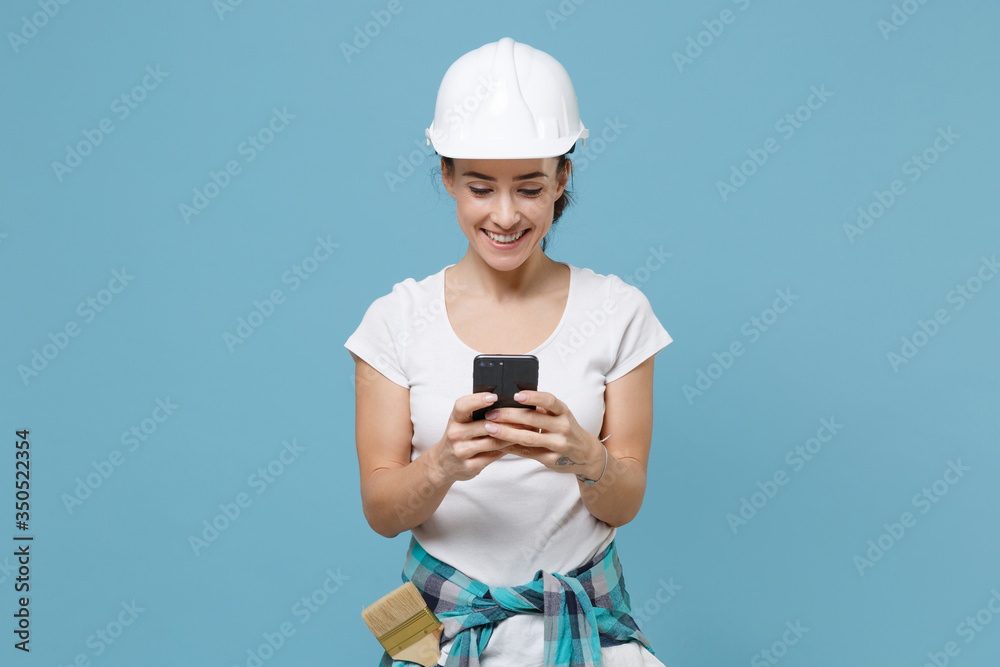 Smiling young woman in protective helmet hardhat using mobile phone, typing sms message isolated on blue background studio. Instruments accessories for renovation apartment room. Repair home concept. Smiling young woman in protective helmet hardhat using mobile phone, typing sms message isolated on blue background studio. Instruments accessories for renovation apartment room. Repair home concept.