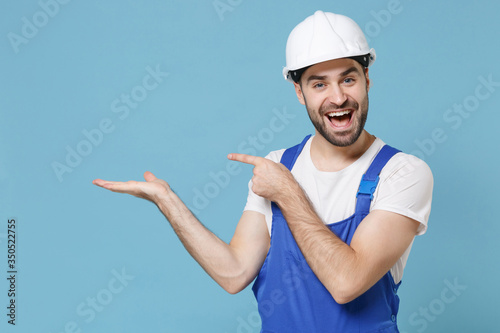 Excited young man in coveralls protective helmet hardhat isolated on blue background. Instruments accessories for renovation apartment room. Repair home concept. Pointing index finger, hand aside.