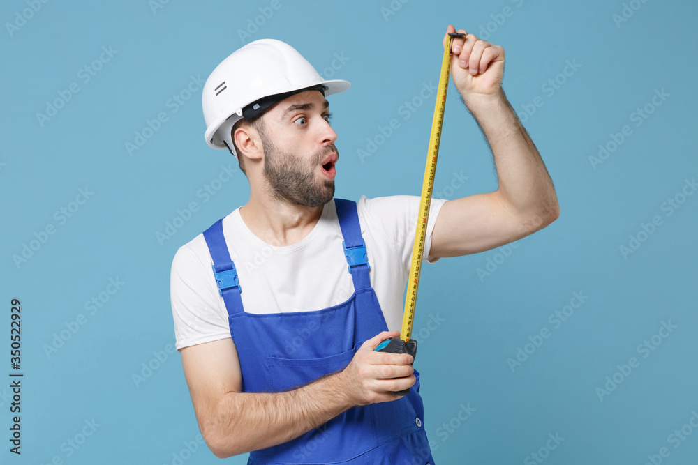 Shocked young man in coveralls protective helmet hardhat hold measure tape isolated on blue wall background studio portrait. Instruments accessories for renovation apartment room. Repair home concept. Shocked young man in coveralls protective helmet hardhat hold measure tape isolated on blue wall background studio portrait. Instruments accessories for renovation apartment room. Repair home concept.