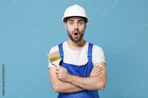 Shocked man in coveralls protective helmet hardhat hold paint brush isolated on blue wall background. Instruments accessories for renovation apartment room. Repair home concept. Holding hands crossed.