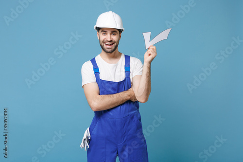 Cheerful young man in coveralls protective helmet hardhat hold check mark isolated on blue wall background studio portrait. Instruments accessories for renovation apartment room. Repair home concept.